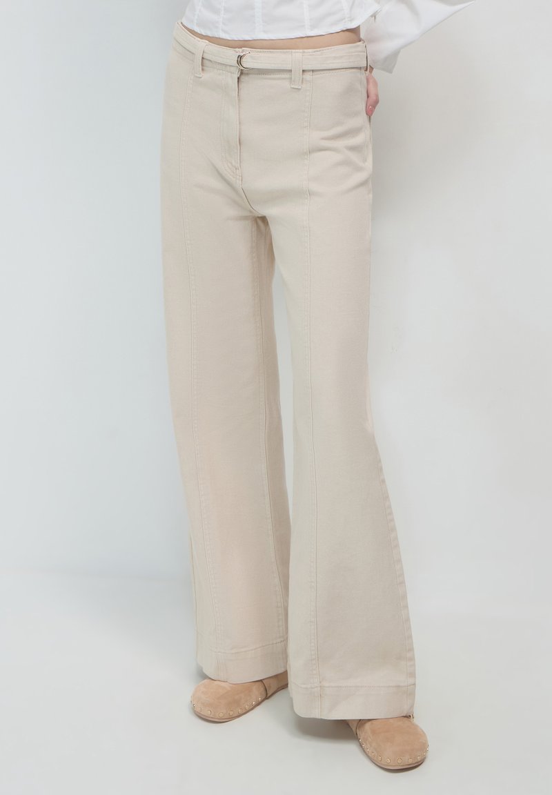 Pantaloni beige a gamba larga realizzati in cotone con cuciture visibili e zip. Presentano vita con passanti per cintura e una vestibilità ampia lungo le gambe.