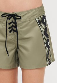 Shorts de bain verts avec un devant lacé noir, présentant une panneautage latérale à motifs floraux et à damier. Texture de tissu lisse.