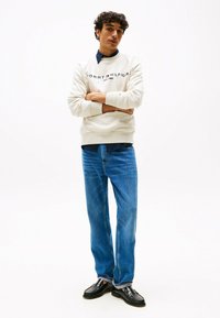 Felpa bianca con logo "TOMMY HILFIGER"; camicia azzurra sotto. Jeans blue straight-leg e scarpe nere completano l'outfit.