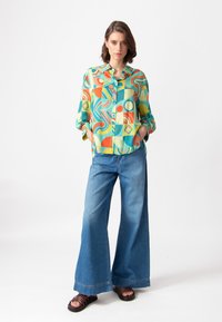 Camisa de botones de colores con patrones abstractos en azul, naranja y verde. Combinada con jeans de mezclilla claros de pierna ancha y sandalias.