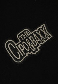 Zwarte stof met een getextureerde, geborduurde merkpatch in het wit, met de tekst "THE CPO & BAXX." Het ontwerp is gedurfd en prominent geplaatst.