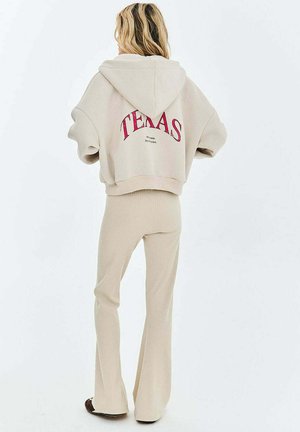 Osoba z długimi włosami, zwrócona tyłem, ubrana w beżową bluzę z kapturem z napisem „TEXAS” na czerwono oraz beżowe prążkowane spodnie na białym tle.