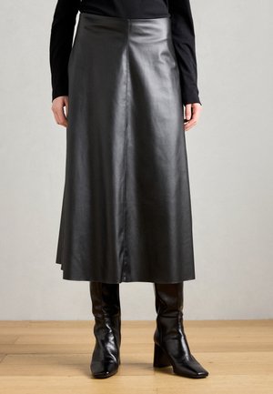 Femme portant une longue jupe en cuir noire, un haut noir à manches longues et des bottes à talons noires, debout sur un sol en bois.