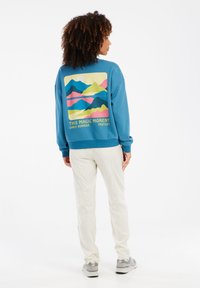 Modrá mikina s okrúhlym výstrihom a grafickým zobrazením horskej scenérie na zadnej strane. Biele nohavice, šedé tenisky a viditeľné kučeravé vlasy.