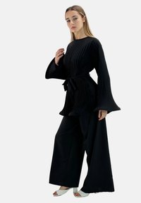 Haut noir plissé avec larges manches et pantalon ample assorti, ceinturé à la taille avec une ceinture, associé à des talons blancs à bout ouvert.