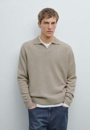 Beige wool sweater with a V-neck and short collar, featuring ribbed cuffs and hem. Model wears it over a white shirt with denim jeans.