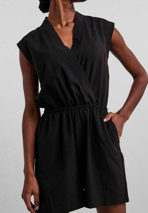 Ærmeløs sort romper med V-udskæring og elastisk talje, båret af en person med hånden i lommen og lys lilla neglelak.