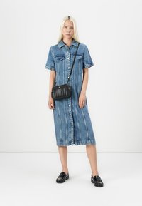 Donna che indossa un vestito midi in denim blu con bottoni e maniche corte, una borsa a tracolla nera e mocassini neri, in piedi davanti a uno sfondo bianco semplice.
