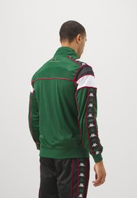 Chaqueta de chándal verde satinado con cuello alto, acentos en negro y blanco, con ribete rojo y patrón de logotipo repetido a lo largo de las mangas.