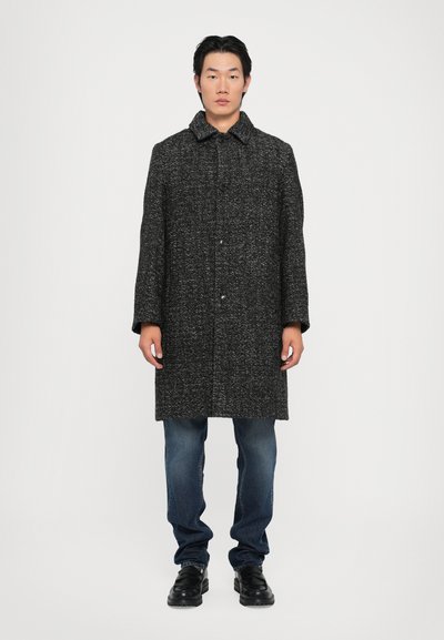 Manteau en mélange de laine noir et gris avec un col, une fermeture à boutons, des poches latérales et une coupe ample, porté sur un jeans bleu et des chaussures noires.