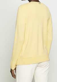 Lys gul sweater med rund hals, ribbede manchetter og kant, samt en subtil tekstur. Bagfra ses et logo-tag nær halsudskæringen.
