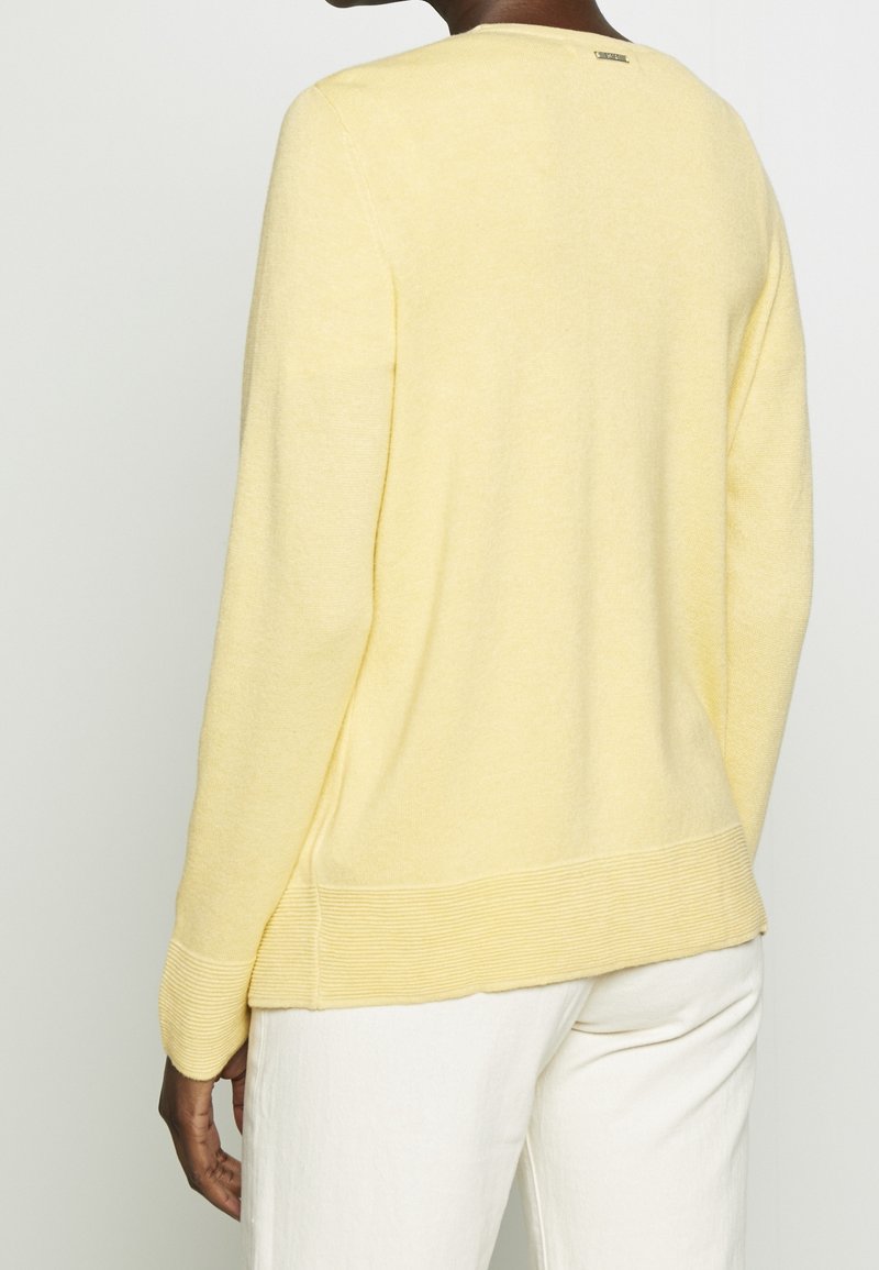 Lys gul sweater med rund hals, ribbede manchetter og kant, samt en subtil tekstur. Bagfra ses et logo-tag nær halsudskæringen.