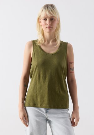 Mladá žena se světlými vlasy, oblečená v olivově zeleném tílku a světle modrých džínách, stojící před jednobarevným bílým pozadím.