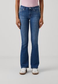 Femme portant un jean bootcut bleu, des baskets blanches et un haut rose sans manches, debout devant un fond clair uni.