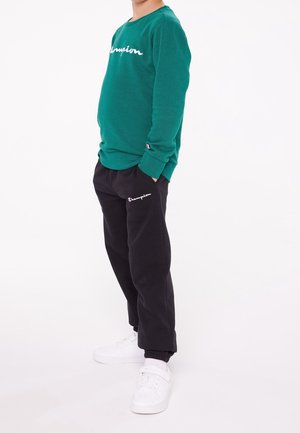 Kind trägt grünes Champion-Sweatshirt und schwarze Jogginghose mit Champion-Logos, steht mit den Händen in den Taschen und weißen Turnschuhen.
