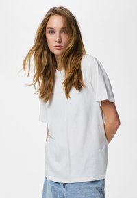 Tânără cu păr lung castaniu deschis purtând un tricou alb simplu cu mâneci scurte și blugi albastru deschis, privind înainte cu mâinile la spate.