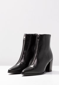 Botas de piel negra hasta el tobillo con cremalleras plateadas en la parte frontal, punteras afiladas y tacones de bloque sobre una superficie y fondo blancos.