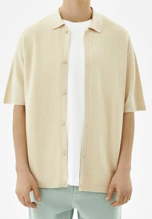 Persoon draagt een beige, gestructureerde korte mouwen cardigan met knopen over een wit T-shirt en lichtblauwe broek, afgebeeld van nek tot heupen.