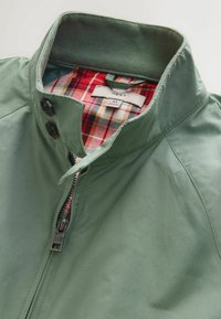 Veste légère verte avec un col montant, une fermeture éclair frontale et deux boutons. L'intérieur présente une doublure à carreaux rouges et noirs.