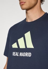 Marineblå bomulds t-shirt med et stort lys_grønt adidas-logo og "REAL MADRID" tekst i hvid, rund hals, korte ærmer, glat tekstur.