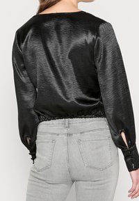Top lungo a maniche nere in satin con orlo arricciato e aperture laterali. Abbinato a jeans aderenti grigio chiaro con tasche posteriori.