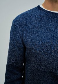 Zemljenost osebe, ki nosi teksturirano temno modro pleteno pulover nad belim majico s krojnim ovratnikom, na eni enotni podlagi.