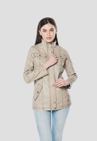 Beige arbejdsjakke med høj krave, fire forlommer, knaplukning foran og kontraststikninger. Stoffet virker blødt med en glat tekstur.