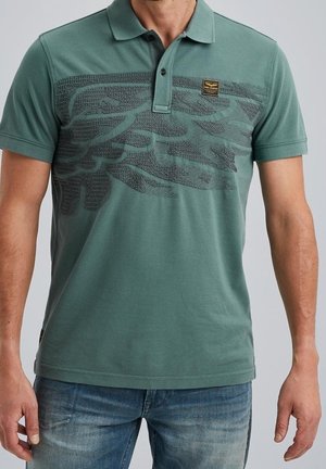 Mężczyzna ubrany w zieloną koszulkę polo z krótkim rękawem i teksturowanym ciemnym wzorem na klatce piersiowej oraz niebieskie dżinsy, stojący na jednolitym tle.