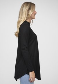 Zwarte top met lange mouwen, hoge hals, losse pasvorm met een gebogen zoom. Zachte stof met een gladde textuur. Gecombineerd met lichtblauwe spijkerbroek.