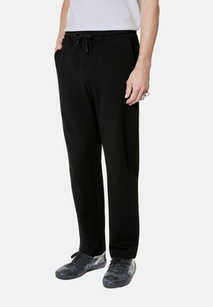 Mann trägt schwarze Sweatpants mit Kordelzug, weißes Hemd, schwarze Turnschuhe, Hände in den Taschen, steht vor weißem Hintergrund.