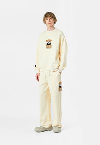 Conjunto de sudadera y pantalones de color crema con diseño de oso de peluche bordado. Tejido suave, corte relajado, y calzado texturizado gris complementan el conjunto.