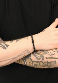 Bracciale di perline nere con un accento cilindrico, indossato su un avambraccio tatuato su uno sfondo nero uniforme. Le perline sono rotonde e opache.