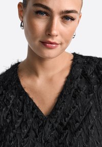 Juodas marškinėliai su V formos iškirpte, puošti tekstūruota fringe detalėmis. Modelis demonstruoja neutralią makiažą ir sidabro spalvos žiedus ausyse prieš šviesų foną.