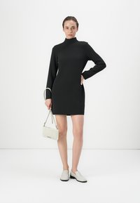 Robe noire à manches longues, col montant, au design ajusté et à la texture lisse ; accompagnée d'un sac à main en tressé blanc et de chaussures claires.