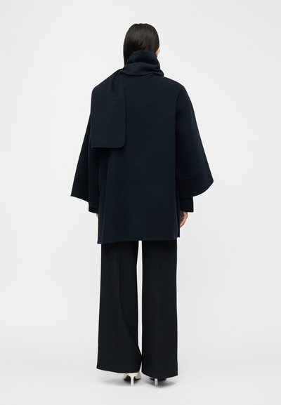 Γυναίκα που φοράει σκουρόχρωμο oversized παλτό με ενσωματωμένη λεπτομέρεια κασκόλ, παντελόνι με φαρδιά μπότα και λευκά τακούνια, στέκεται με την πλάτη προς το φακό σε λευκό φόντο.