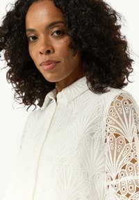 Weiße bestickte Bluse mit kragenähnlichem Ausschnitt und detaillierten Spitzenärmeln, die ein Pfauenfeder-Muster und strukturierten Stoff aufweist.