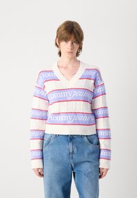 Witte kabelgebreide trui met V-hals, met horizontale banden in lichtblauw en roze met de tekst "tommy jeans". Draagt met blauwe jeans.