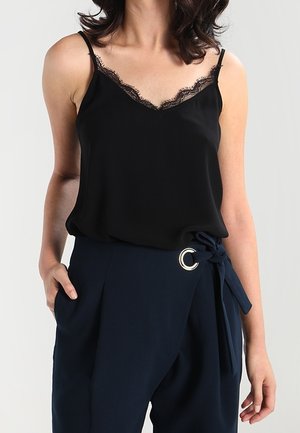 Kvinde iført en sort camisole med blonder, der er stoppet ned i navy omslagsbukser med bindebælte og sølvringdetalje.