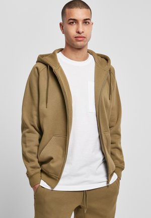 Tamsiai žalia zip-up gobtuvė su priekinėmis kišenėmis, derinama su balta marškinėliais. Medžiaga yra minkšta, lygi ir kasdieniška.