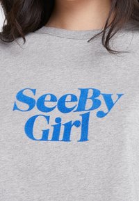 Camiseta de algodón gris con un audaz texto gráfico azul "SeeBy Girl" en una tipografía llamativa. De manga corta y con un clásico escote redondo.
