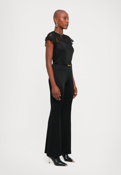 Femme avec la tête rasée portant un haut à manches en dentelle noire, un pantalon flare taille haute avec une ceinture, des boucles d'oreilles en or, et des escarpins noirs à bouts en argent.