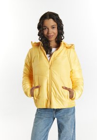 Giacca a vento gialla con zip frontale, cappuccio e polsini elasticizzati. Presenta una texture liscia e lucida e due tasche frontali. Abbinata a jeans blu.