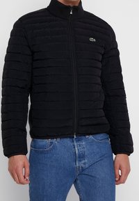 Man med en svart uppblåsbar jacka med hög krage, ihop med blå jeans, stående mot en enfärgad bakgrund.