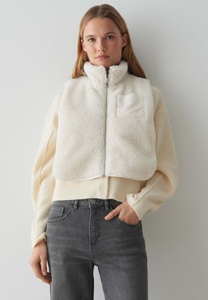 Kurzes Weste aus Kunstfell in Off-White mit hohem Kragen, Reißverschluss vorne und einer Brusttasche. Getragen über einem hellen Cremepullover und grauen Jeans.
