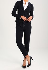 Marinblått rutig kostym med en figursydd kavaj och avsmalnande byxor, tillverkad av mjukt tyg. Matchad med svarta högklackade skor och en vit skjorta.