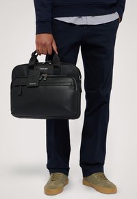 Svart läder portfölj med dubbla dragkedjor, en framficka och ett bagagetag. Har slitstarka handtag och en slät yta. Designen är rektangulär.