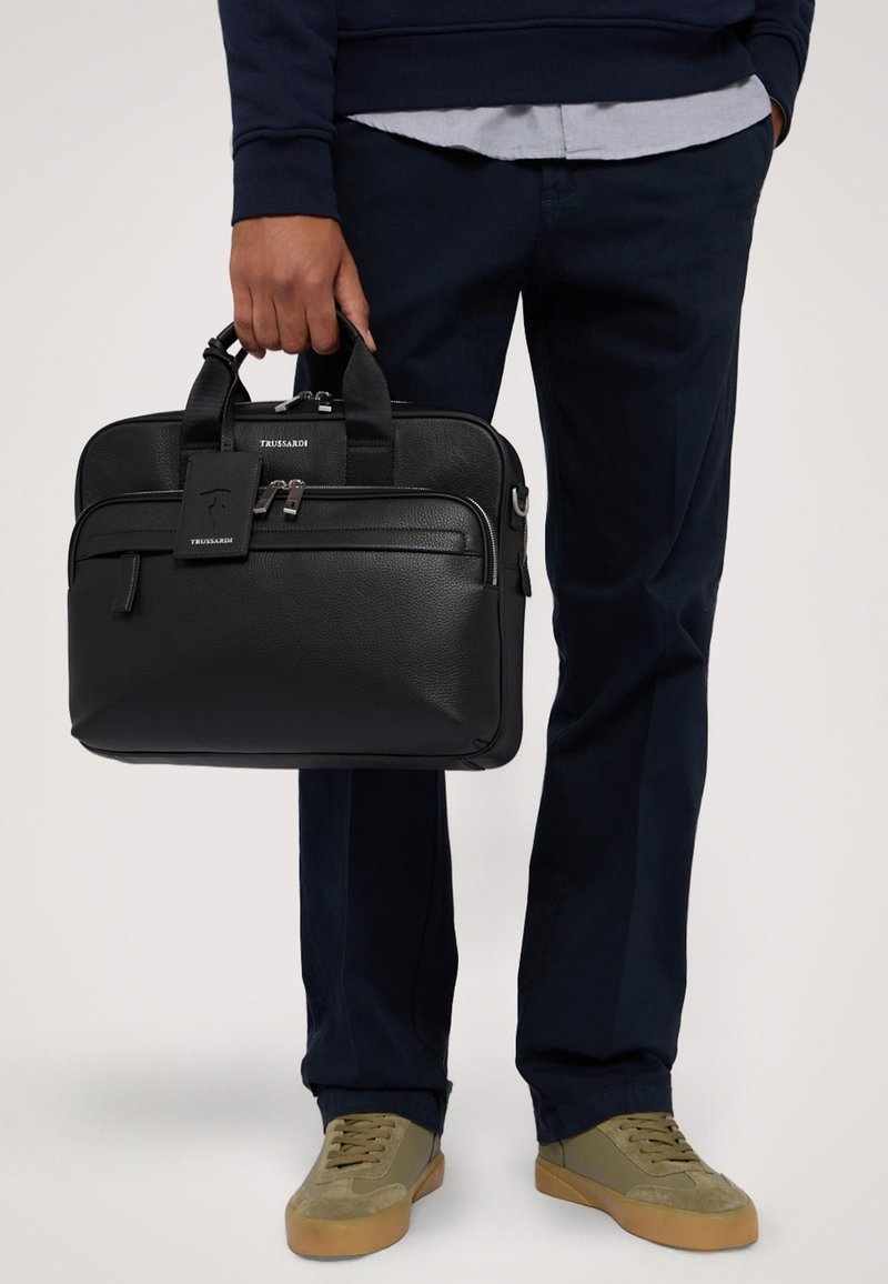 Svart läder portfölj med dubbla dragkedjor, en framficka och ett bagagetag. Har slitstarka handtag och en slät yta. Designen är rektangulär.