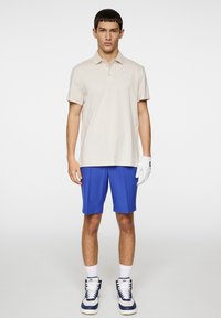 Beige pikétröja, blåa shorts, vita strumpor och blå sneakers. Outfiten har en klassisk design med krage och diskret branding.