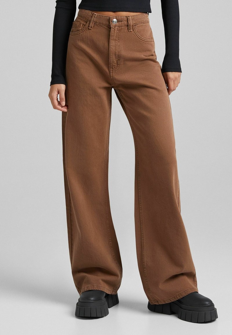 Personne portant un pantalon marron taille haute à jambes larges avec des poches avant, associé à des chaussures plateformes noires épaisses et un haut noir à manches longues.