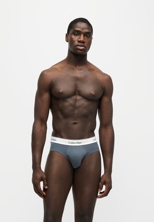 Spierbundige man staat tegen een witte achtergrond, hij draagt blauwe Calvin Klein slips met een witte tailleband.